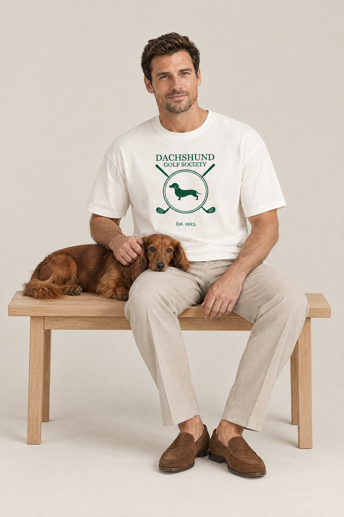 Man sitting on a bench with a long-haired dachshund in the white Dachshund Golf Society Tee. Color: White