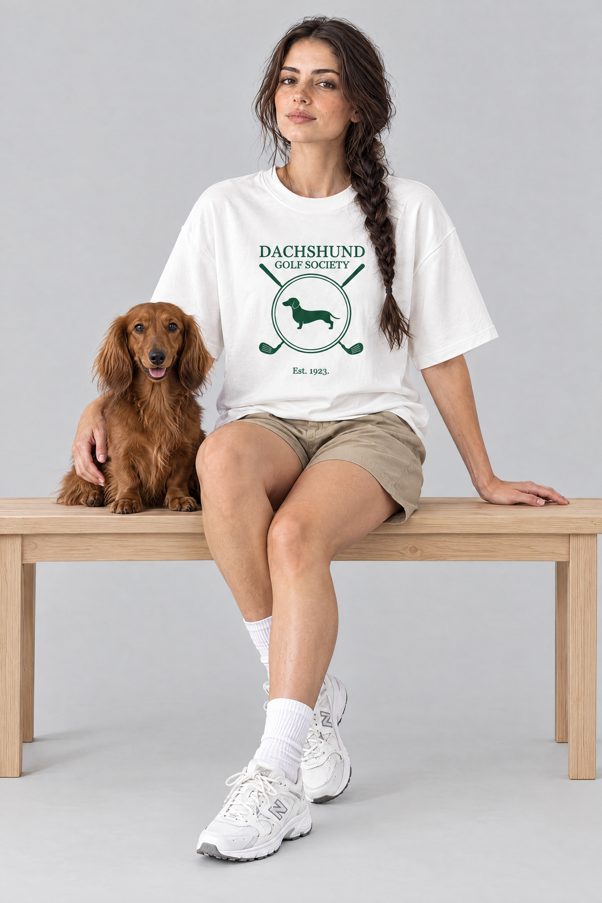 Woman sitting on a bench with a long-haired dachshund in the white Dachshund Golf Society Tee. Color: White