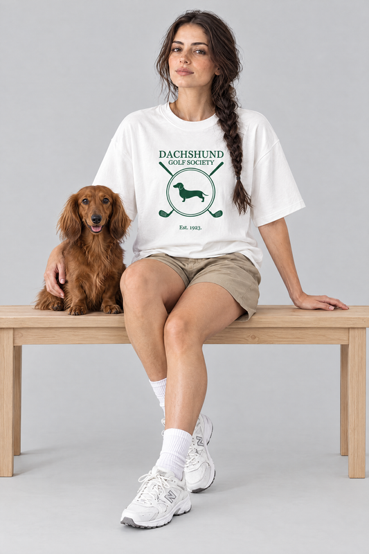 Woman sitting on a bench with a long-haired dachshund in the white Dachshund Golf Society Tee. Color: White