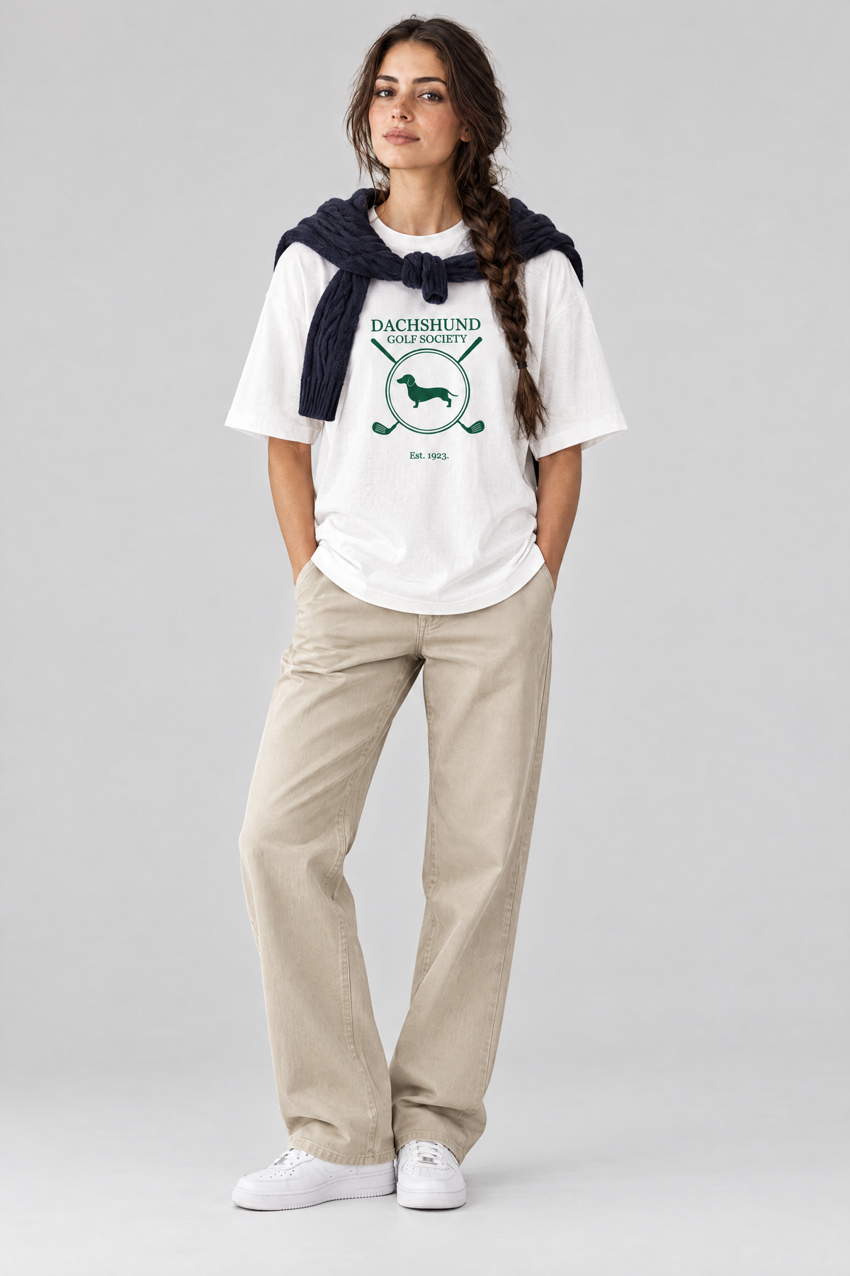 Woman wearing the Dachshund Golf Society Tee in white with stone trousers and a navy sweater over her shoulders. Color: White