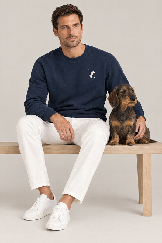 Man sitting on a wooden bench with a wire-haired dachshund in the navy Swing dachshund golf sweatshirt and white chinos. Color: Navy