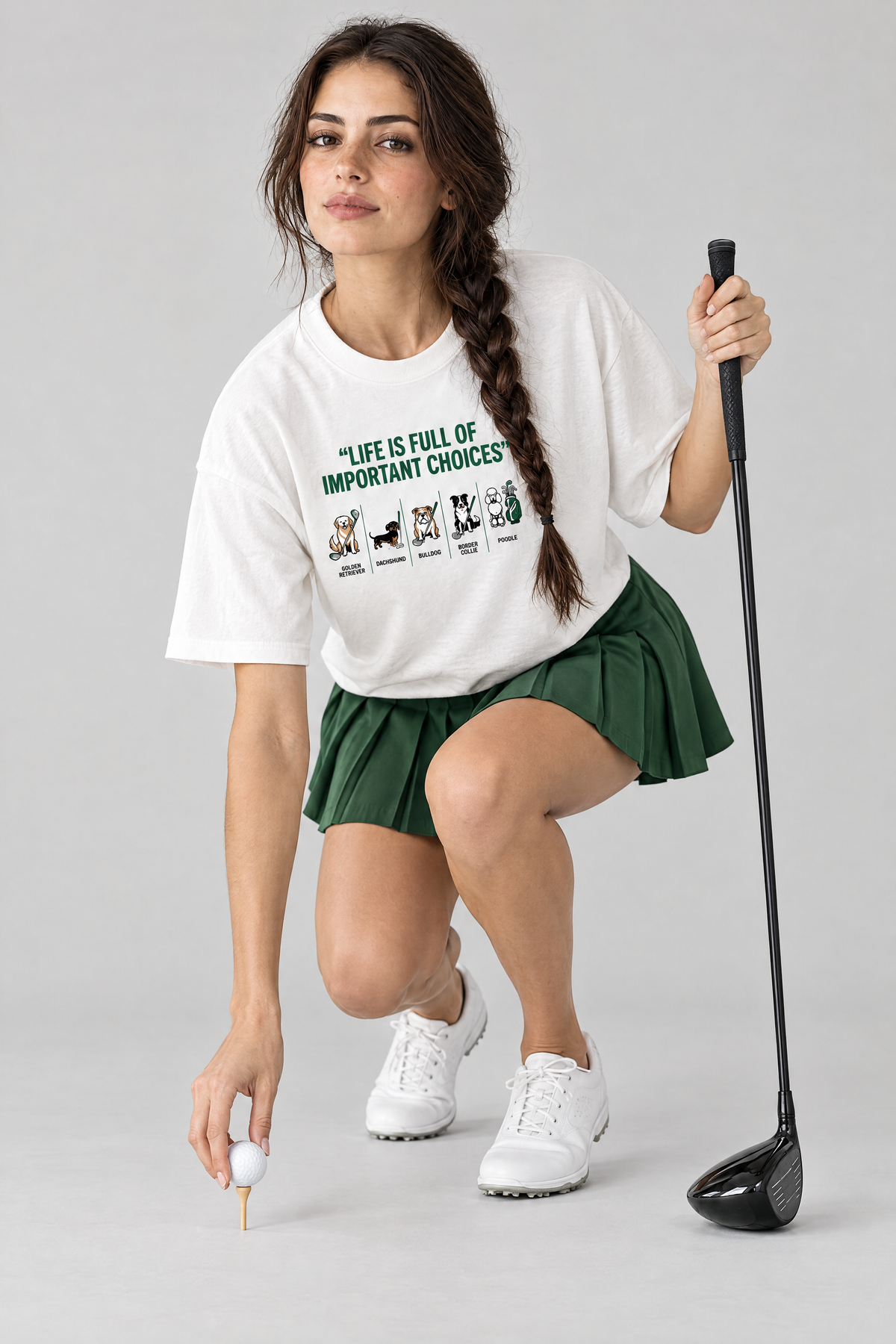 Woman teeing up a golf ball in the white Full of Choices Dog Golf Tee and a green pleated skirt. Color: White