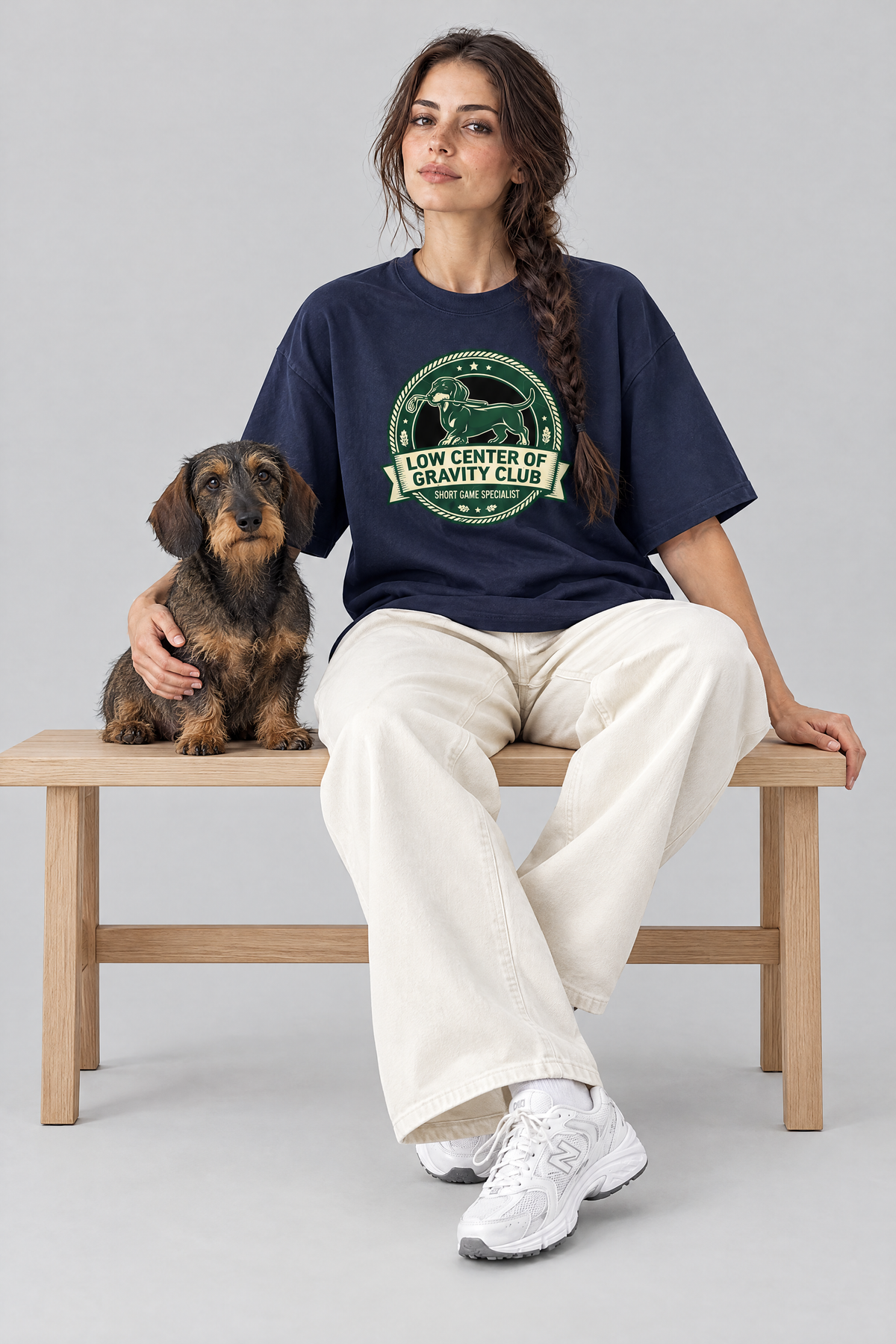 Woman sitting on a bench with a wire-haired dachshund in the navy Low Center of Gravity Club Dachshund Golf Tee. Color: Navy