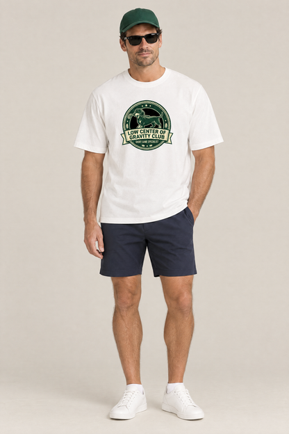 Man wearing the Low Center of Gravity Club Dachshund Golf Tee in white with navy golf shorts, a green cap and sunglasses. Color: White