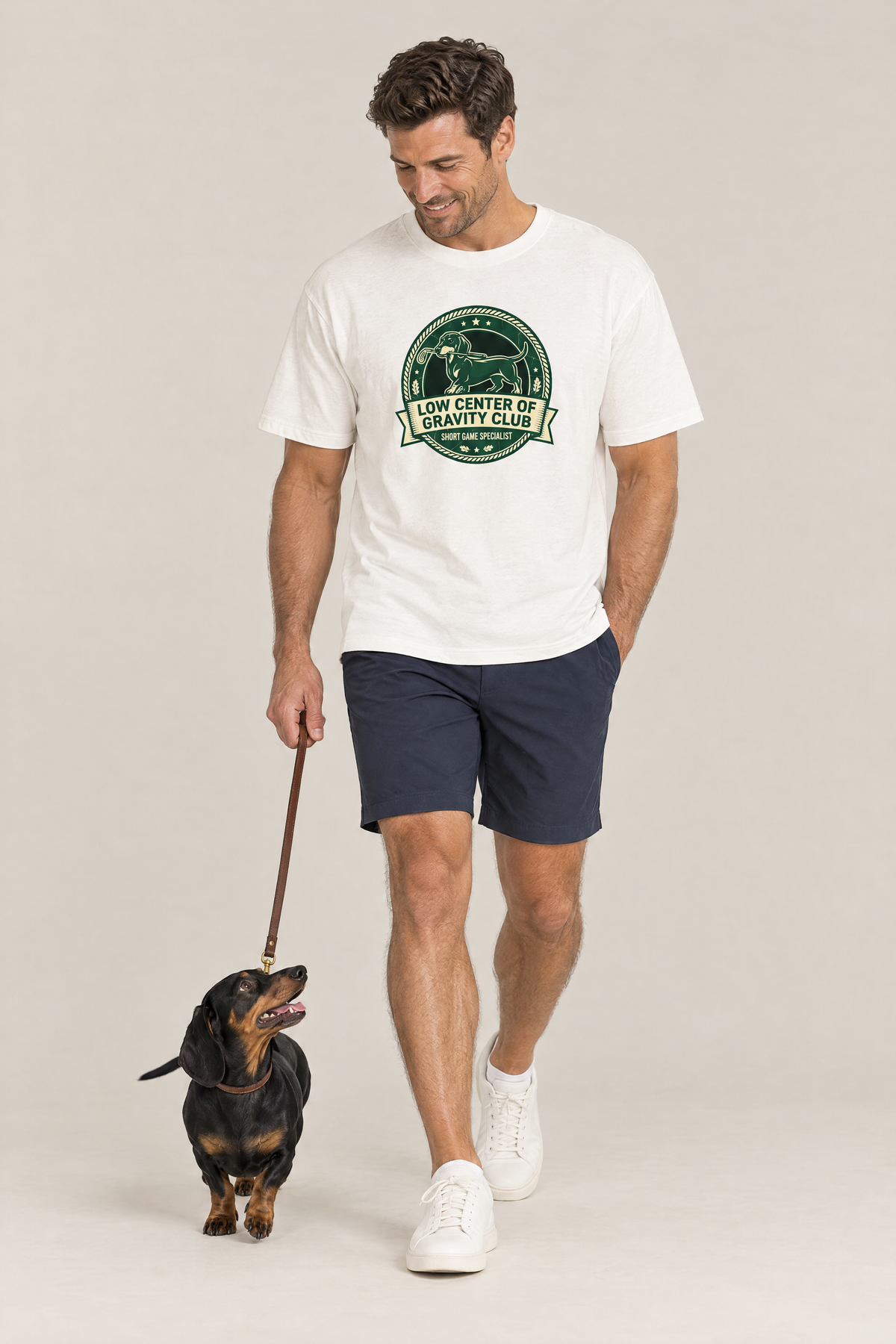 Man walking a dachshund on a leash in the white Low Center of Gravity Club Dachshund Golf Tee. Color: White