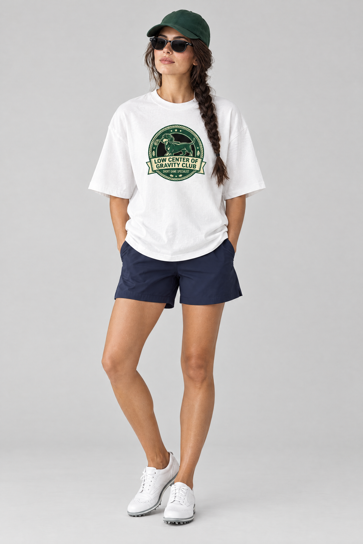 Woman wearing the Low Center of Gravity Club Dachshund Golf Tee in white with navy golf shorts, a green cap and sunglasses. Color: White
