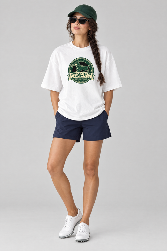 Woman wearing the Low Center of Gravity Club Dachshund Golf Tee in white with navy golf shorts, a green cap and sunglasses. Color: White