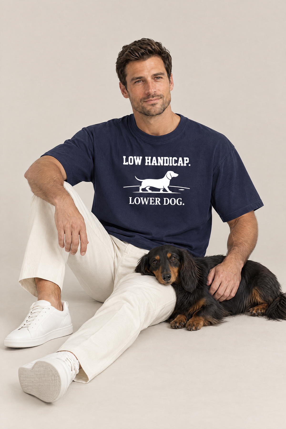 Man sitting on the floor with a long-haired dachshund in the navy Low Handicap Dachshund Golf Tee. Color: Navy