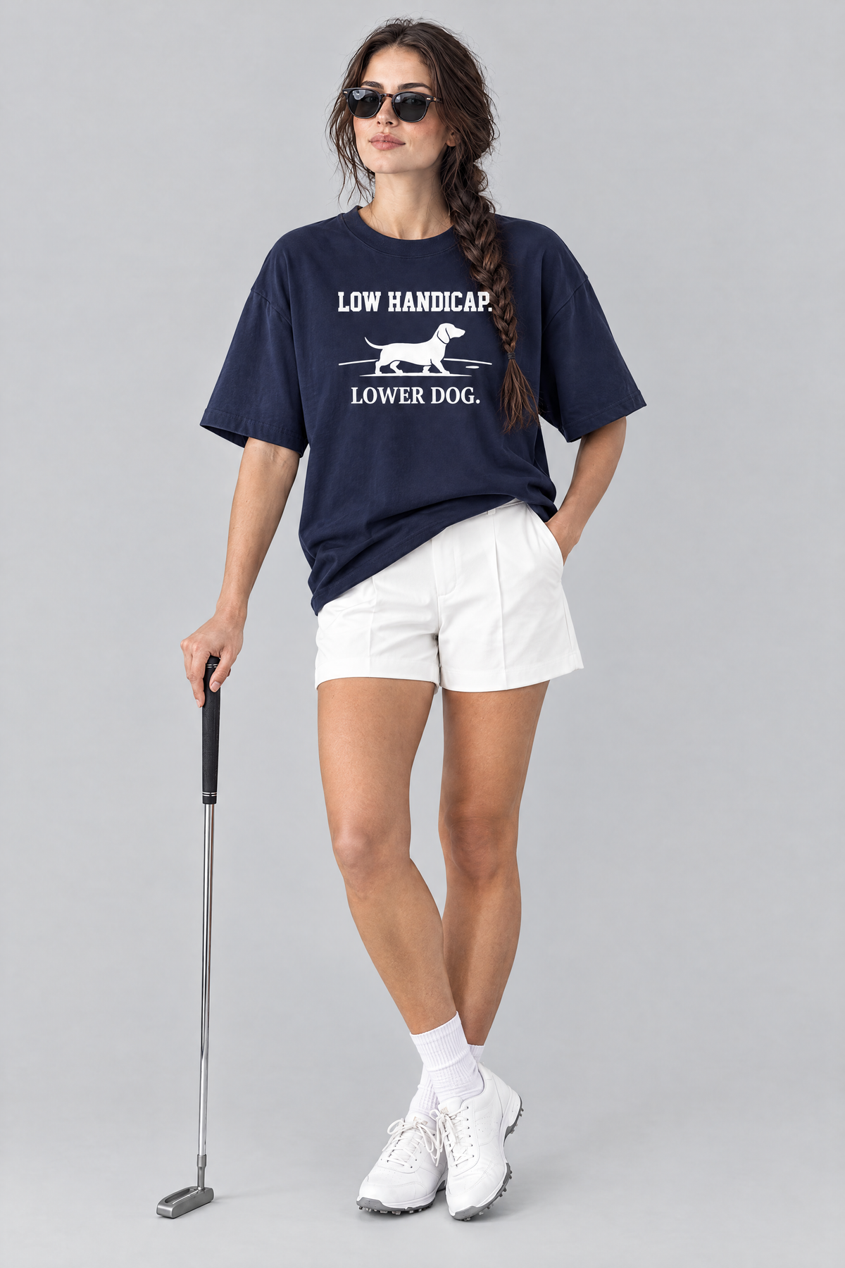 Woman wearing the Low Handicap Dachshund Golf Tee in navy with white tailored shorts and sunglasses, holding a putter. Color: Navy