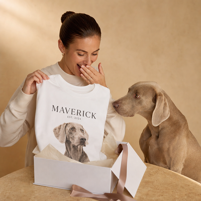Woman holding a white sweatshirt with a dog's photo and name, standing next to a dog on a beige background.