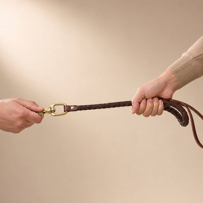 Two hands pulling on a brown leather strap with a gold buckle against a beige background