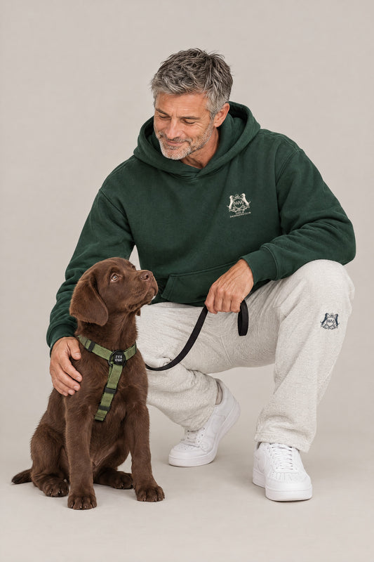 Man in the blackish green Club Crest hoodie and oat Club Crest sweatpants crouching with a Labrador puppy in the green Cloud 7 RESC7UE harness