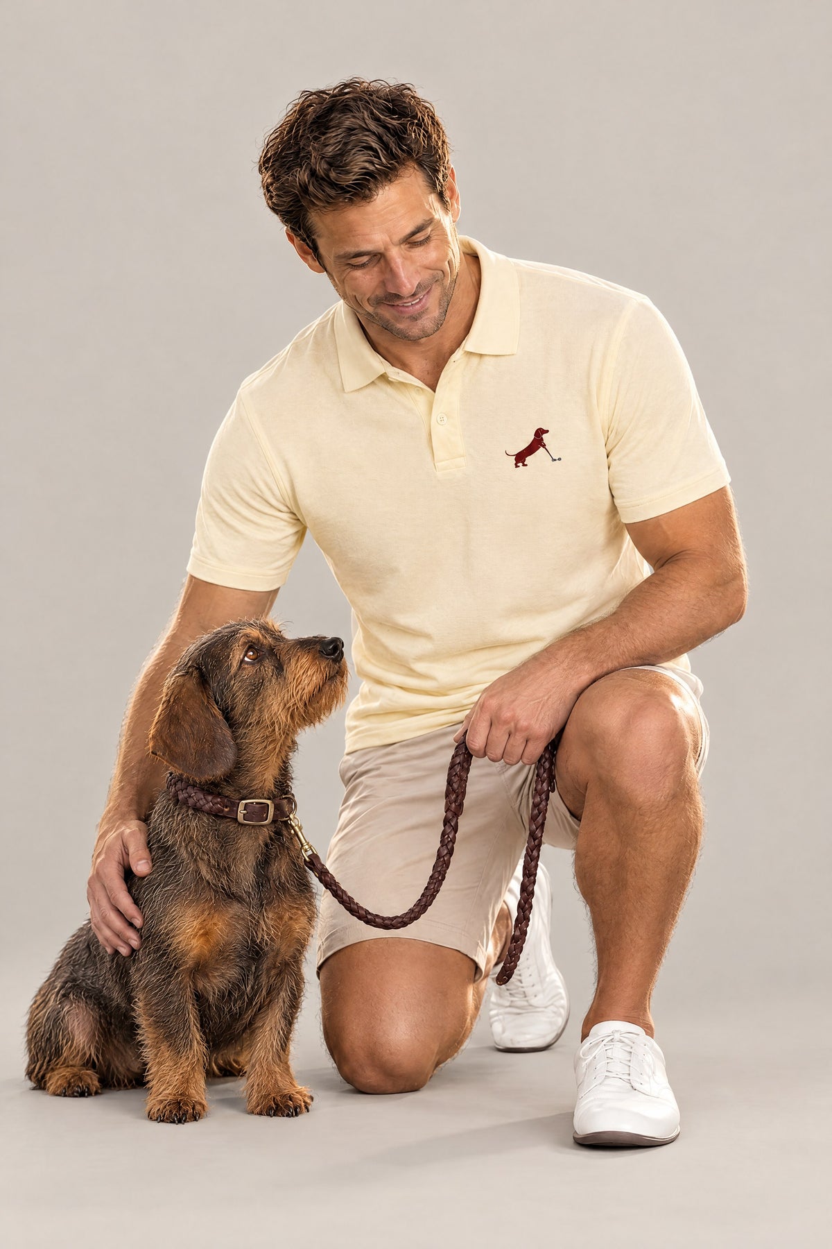 Man in the butter yellow Putting dachshund golf polo kneeling next to a wire-haired dachshund in the saddle brown Cloud 7 Central Park collar