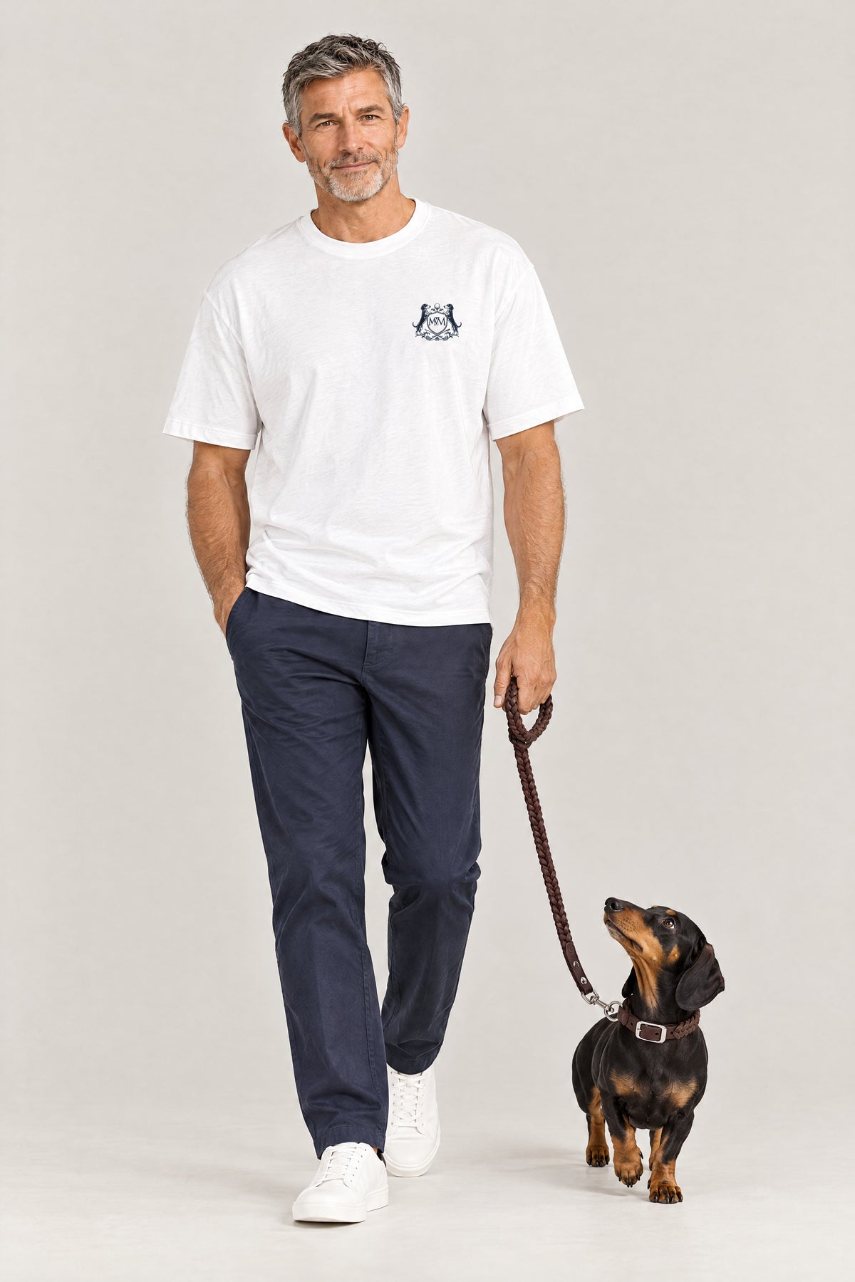 Man in the white Club Crest tee walking a dachshund wearing the brown Cloud 7 Central Park collar and braided leash