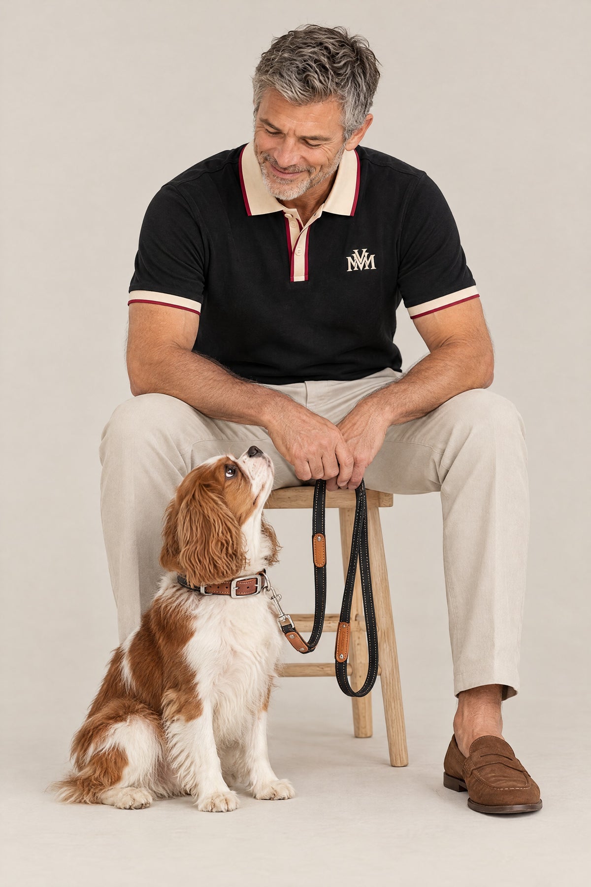 Man in the black Club Crest color block polo on a stool with a Cavalier King Charles Spaniel wearing the Cloud 7 Tivoli Black collar