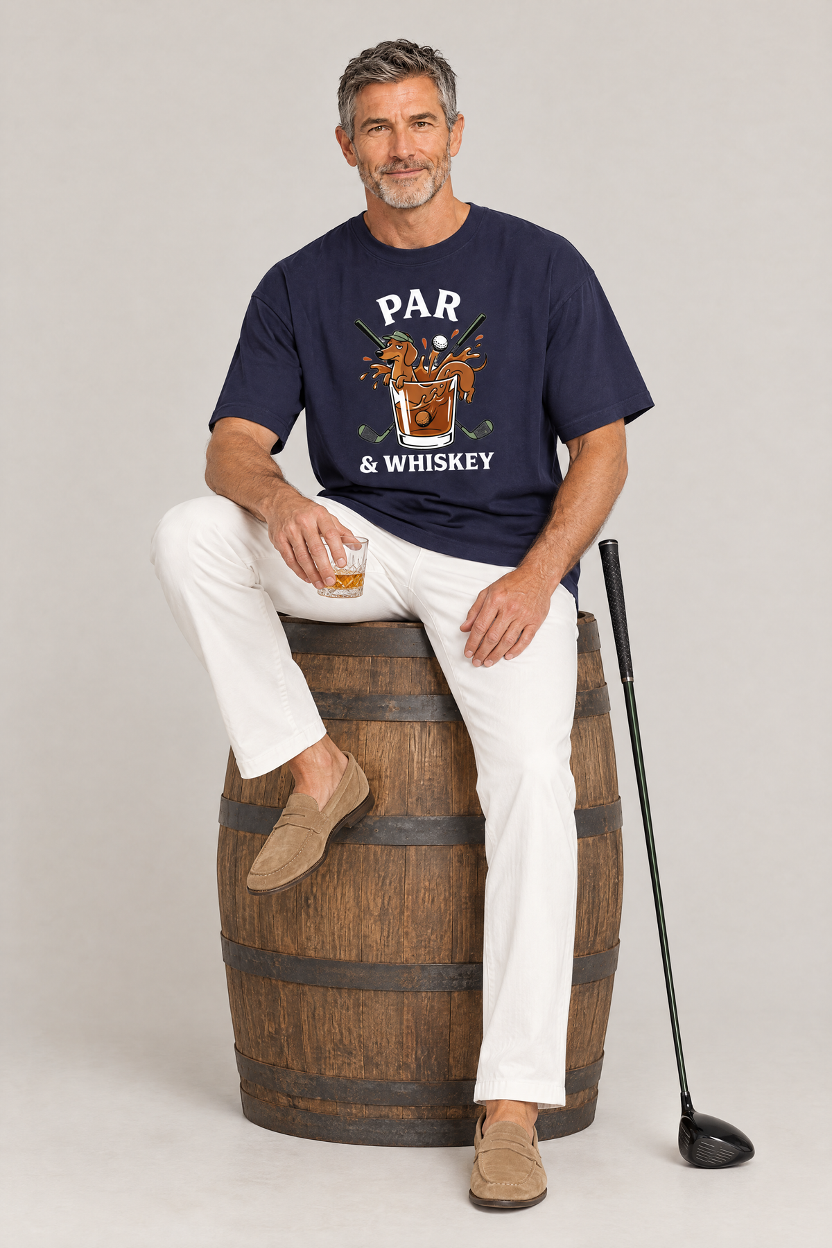 Man sitting on a whisky barrel with a golf club in the navy Par & Whiskey Dachshund Golf Tee. Color: Navy