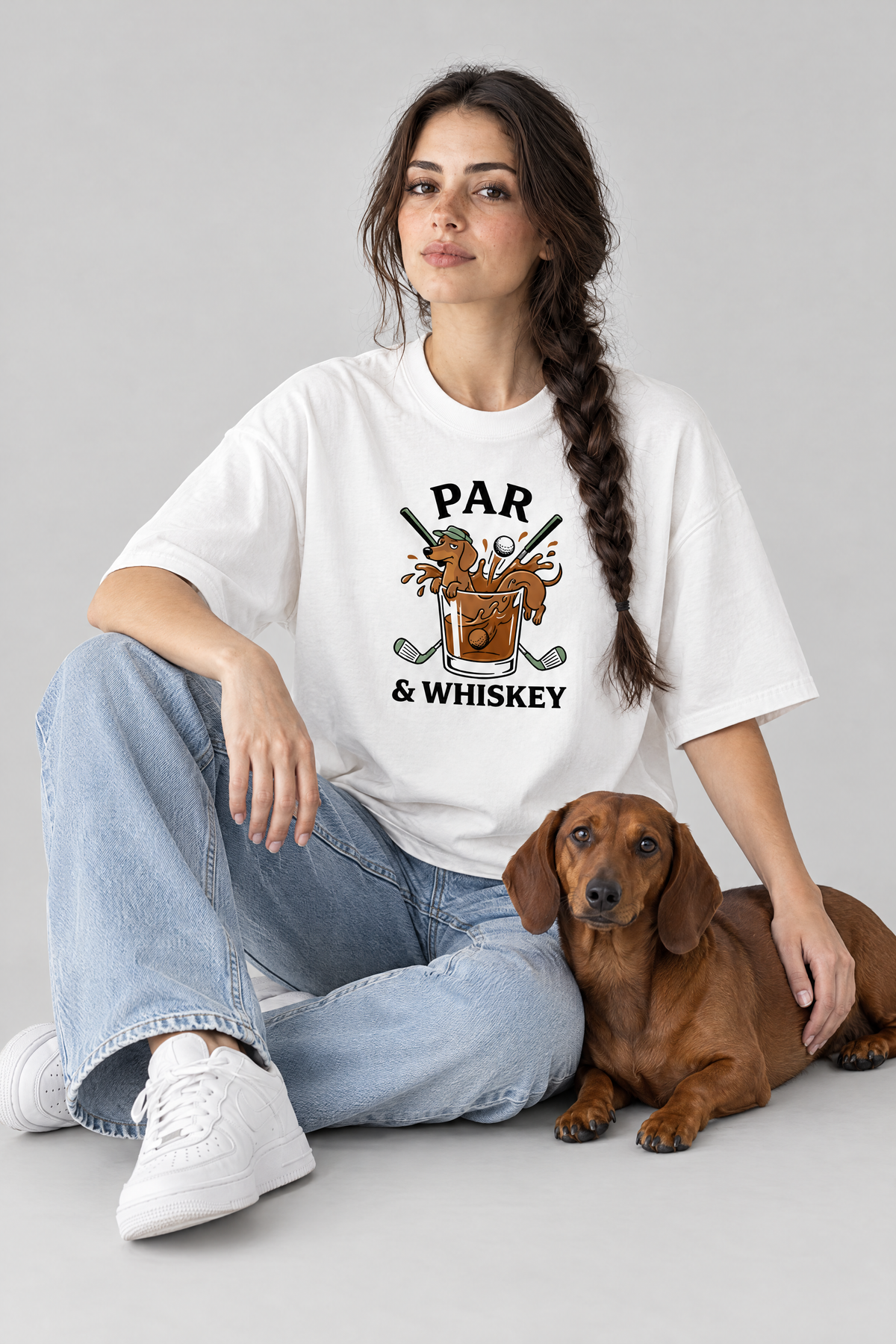 Woman sitting on the floor with a red dachshund in the white Par & Whiskey Dachshund Golf Tee. Color: White
