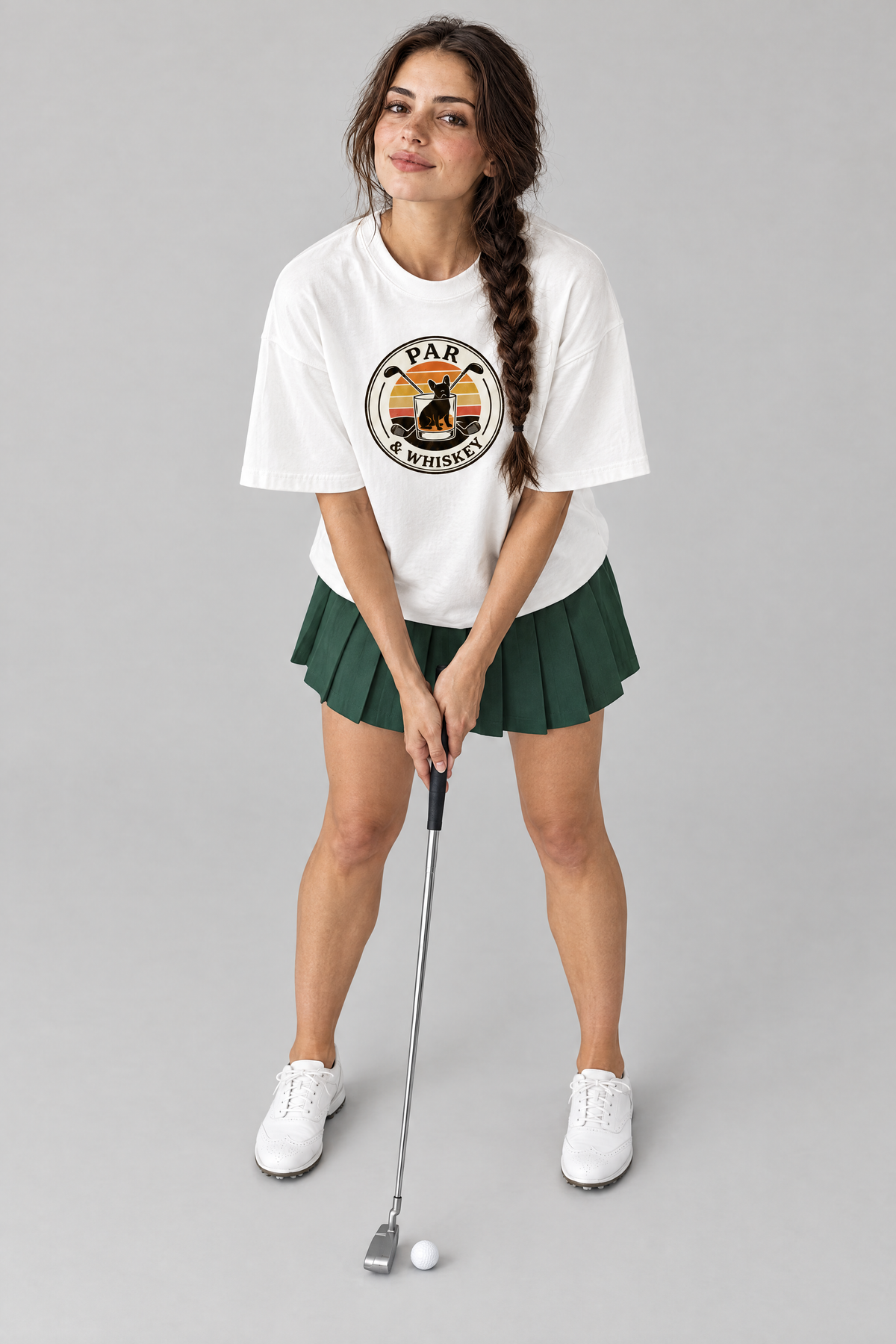 Woman lining up a putt in the white Par & Whiskey French Bulldog Badge Golf Tee and a green pleated skirt. Color: White