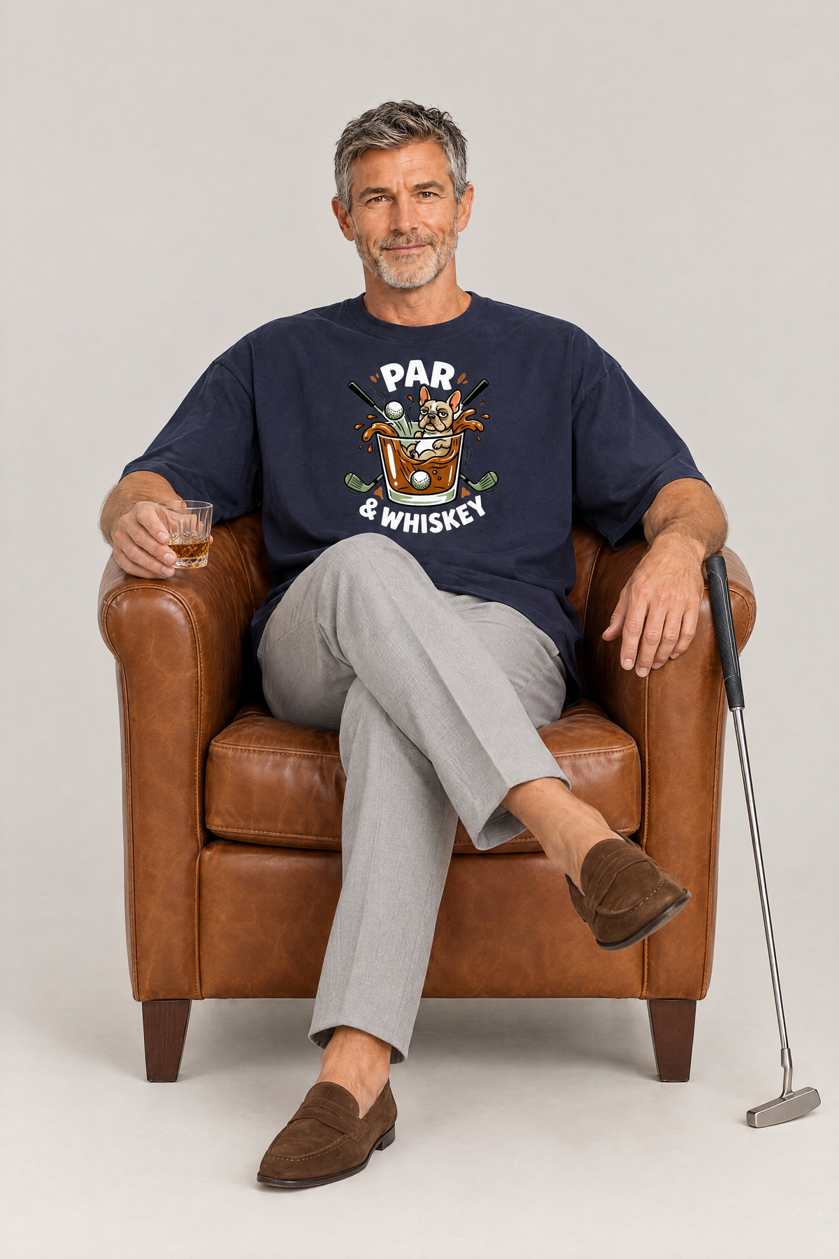 Man sitting in a leather armchair with a whisky glass in the navy Par & Whiskey French Bulldog Splash Golf Tee. Color: Navy