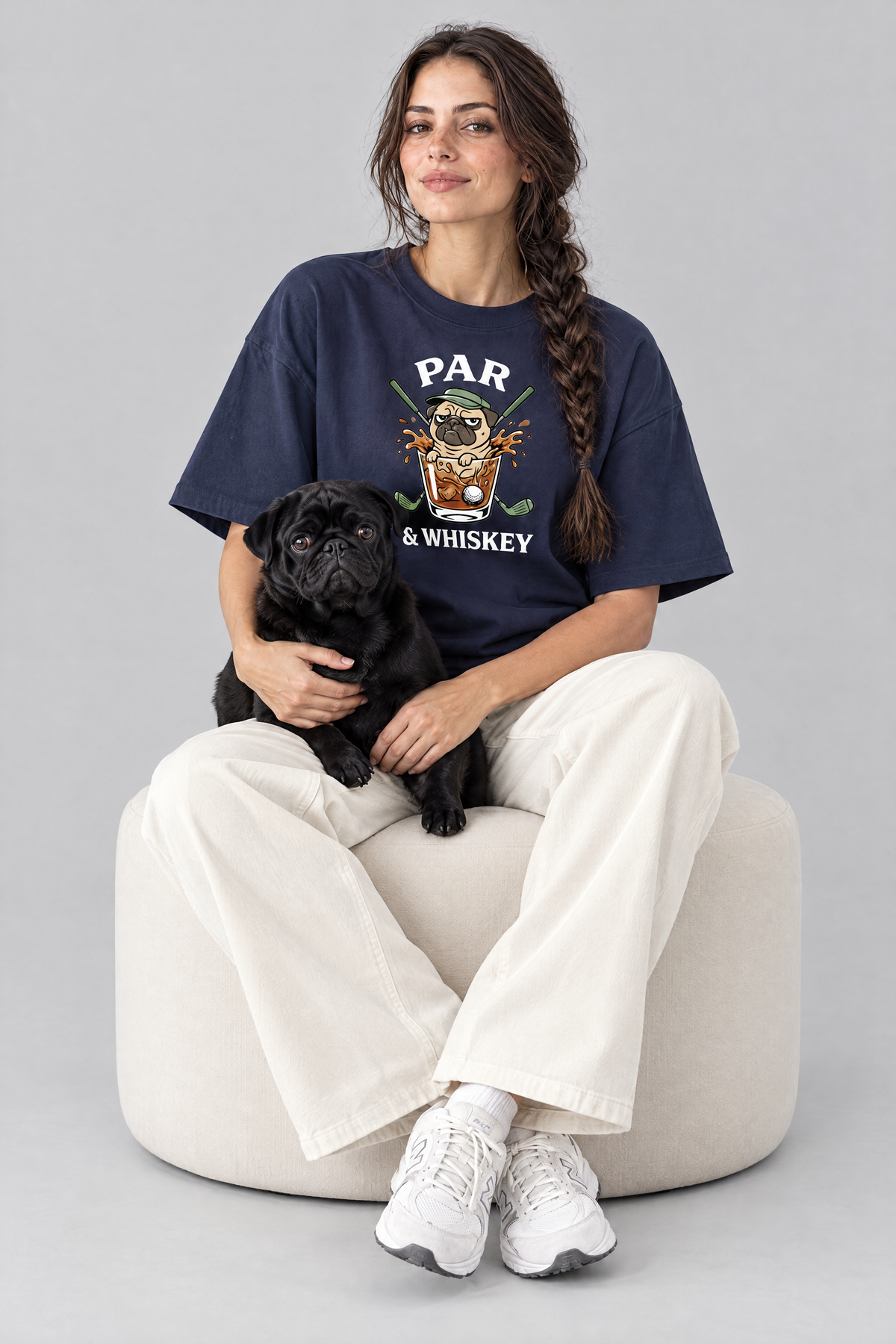 Woman sitting on an ottoman with a black pug in the navy Par & Whiskey Pug Golf Tee. Color: Navy
