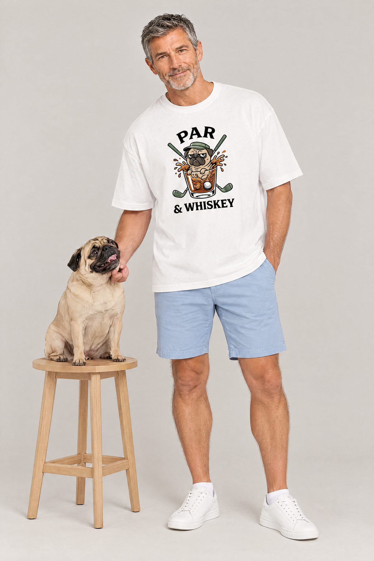 Man wearing the Par & Whiskey Pug Golf Tee in white with a pug on a wooden stool. Color: White