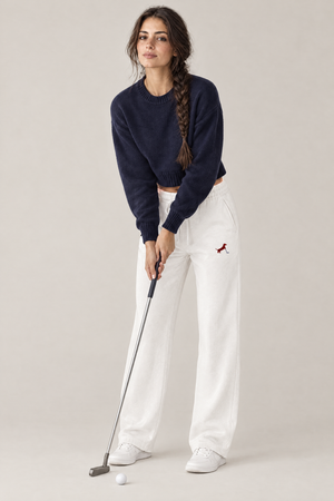 Woman in a relaxed putting stance with a putter in white Putting dachshund golf sweatpants with the small burgundy dachshund putter print on the thigh and a navy cropped sweater. Color: White