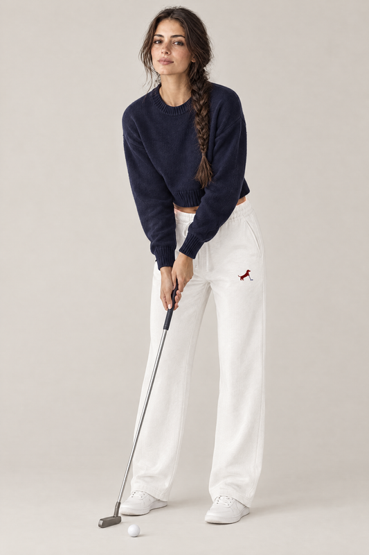 Woman in a relaxed putting stance with a putter in white Putting dachshund golf sweatpants with the small burgundy dachshund putter print on the thigh and a navy cropped sweater. Color: White