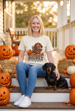 Frau im Dackel Halloween T-Shirt in Natural mit Dackel neben einer zerbrochenen Kürbislaterne und dem Spruch Regret Was Never An Option, auf einer Herbstveranda mit einem schwarz-roten Dackel und Kürbissen