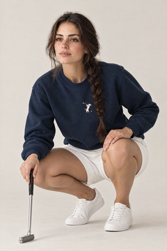 Woman crouching in a golfer putt-reading pose wearing navy Swing dachshund golf sweatshirt and white shorts. Color: Navy