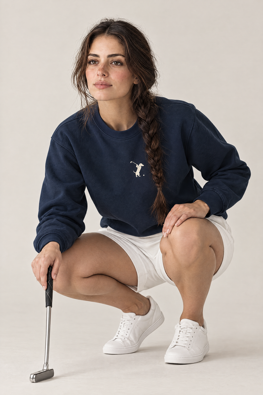 Woman crouching in a golfer putt-reading pose wearing navy Swing dachshund golf sweatshirt and white shorts. Color: Navy