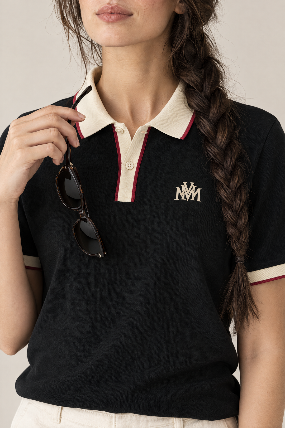 Woman holding sunglasses at the collar of the black Club Crest polo, close-up of the apricot monogram. Color: Black with Apricot