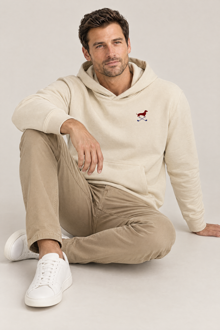 Smiling man sitting on the studio floor in apricot Club & Swing dachshund golf hoodie and khaki chinos. Color: Apricot