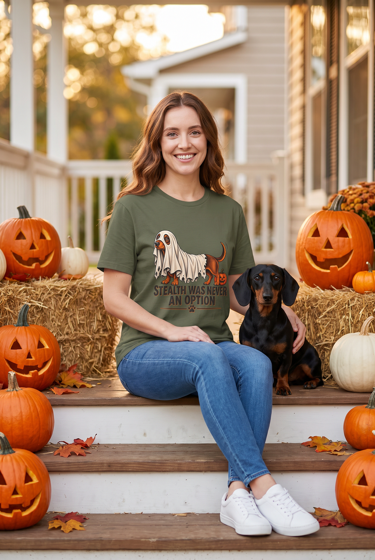 Frau im Dackel Halloween T-Shirt in Military Green mit Dackel unter einem zu kurzen Gespensterlaken und dem Spruch Stealth Was Never An Option, auf einer Herbstveranda mit einem schwarz-roten Dackel und Kürbissen