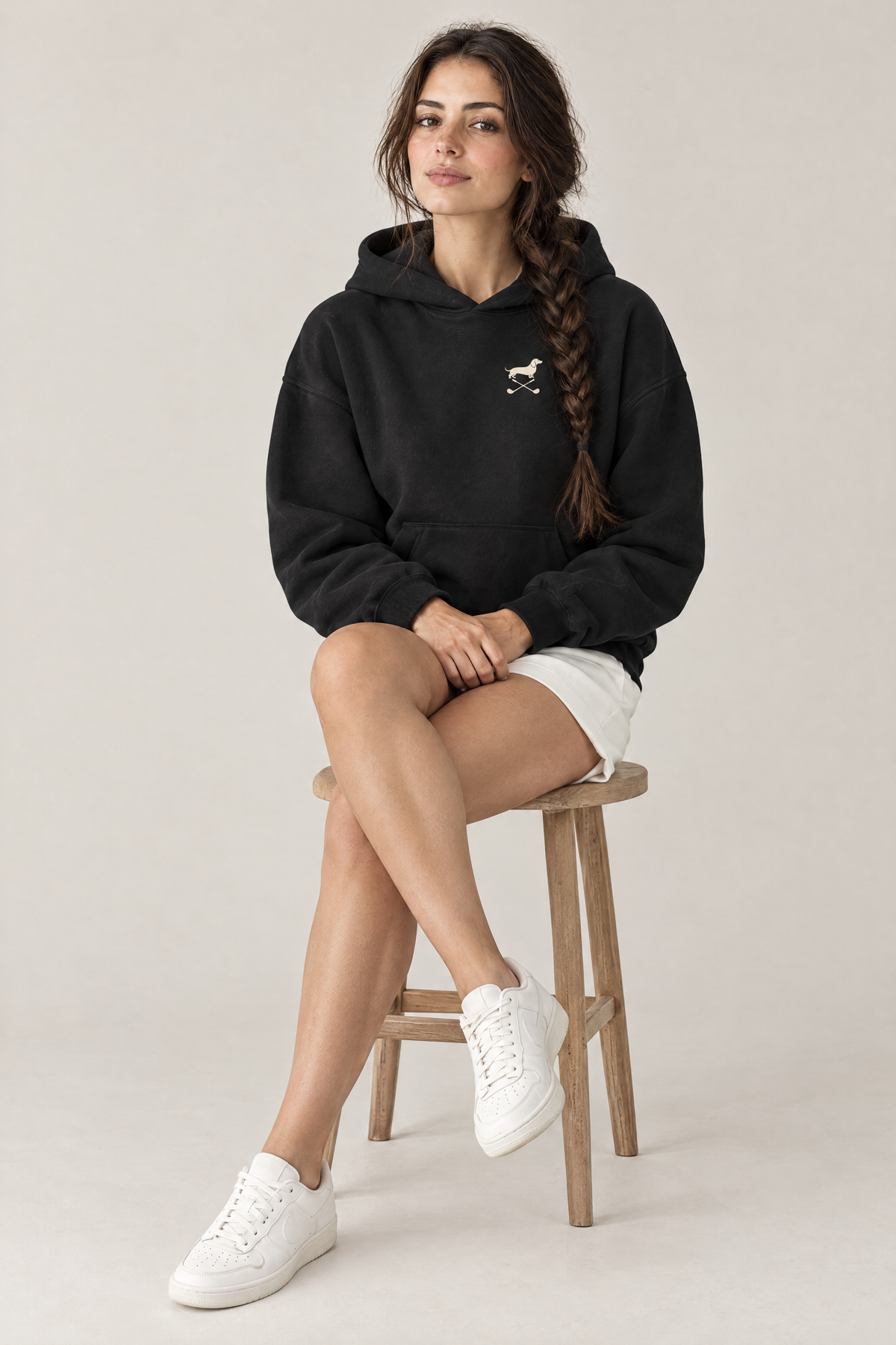 Woman sitting on a wooden stool wearing black Club & Swing dachshund golf hoodie with white shorts. Color: Black