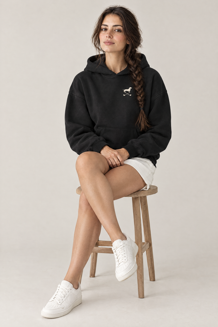 Woman sitting on a wooden stool wearing black Club & Swing dachshund golf hoodie with white shorts. Color: Black