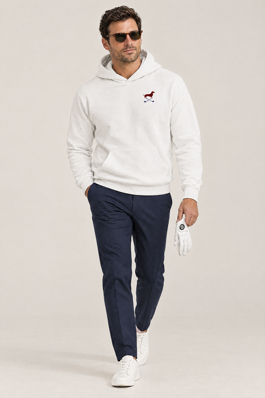Man in dark sunglasses walking in white Club & Swing dachshund golf hoodie and navy trousers, holding a golf glove, full-body studio look. Color: White