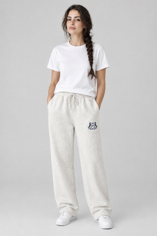 Woman in oat gray Club Crest sweatpants with the small navy crest on the thigh and a white tee, full body. Color: Oat Gray