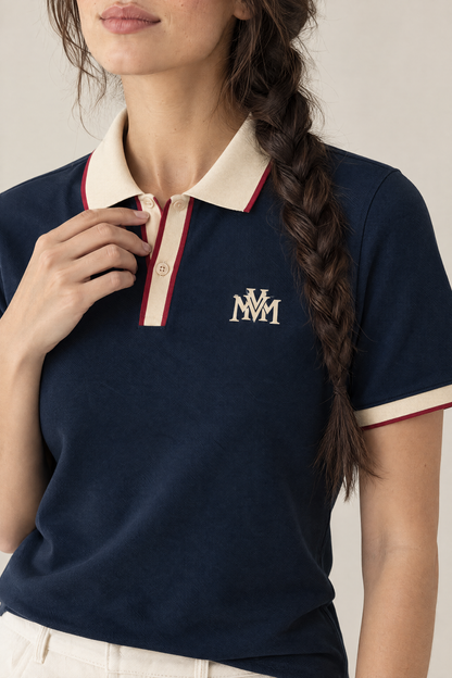 Woman touching the apricot placket of the navy Club Crest polo, close-up of the MVM monogram. Color: Navy Blue with Apricot