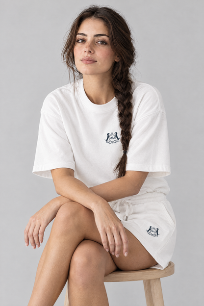Woman sitting on a wooden stool in white Club Crest shorts with the small navy crest on the upper thigh. Color: White