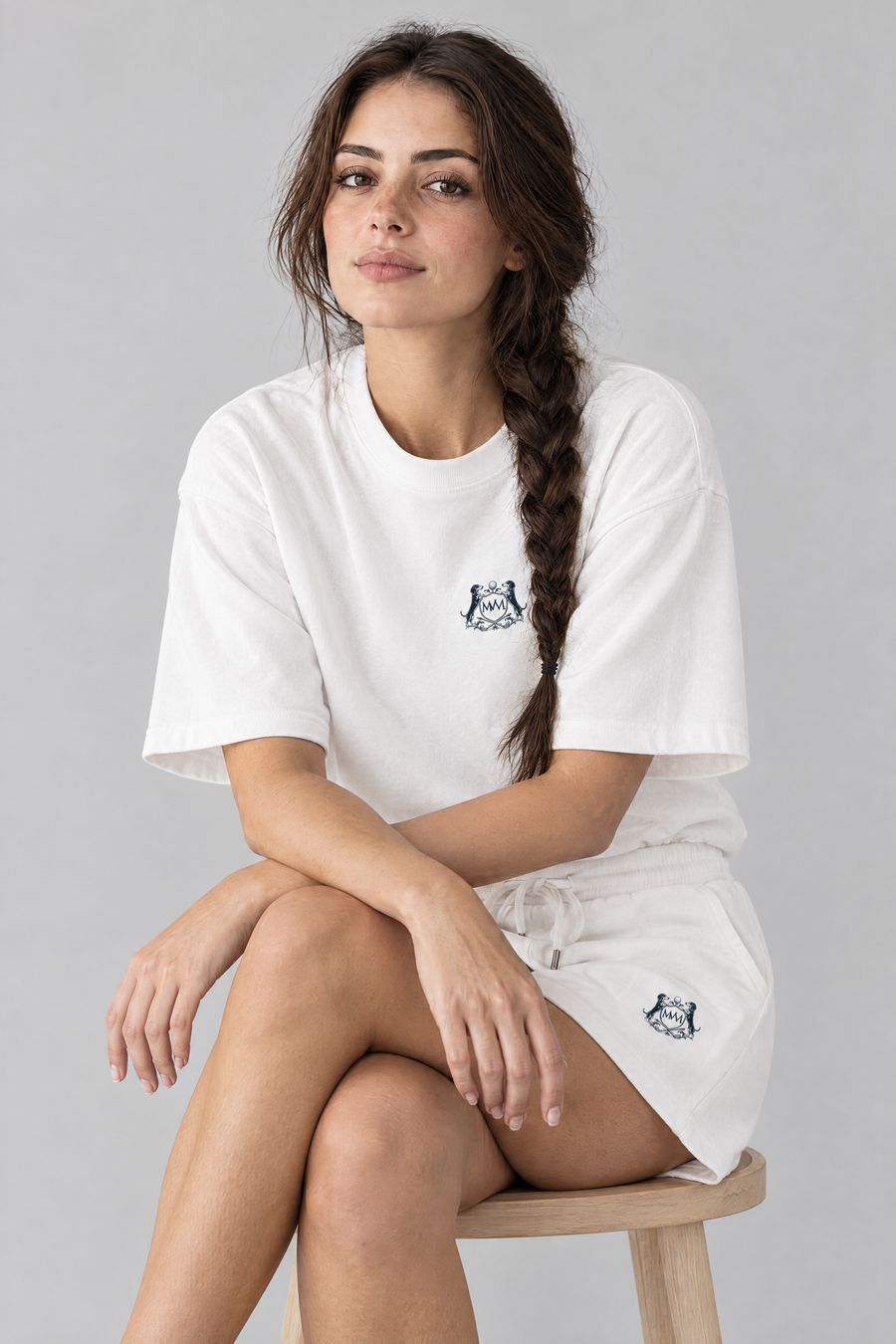 Woman sitting on a wooden stool in white Club Crest shorts with the small navy crest on the upper thigh. Color: White