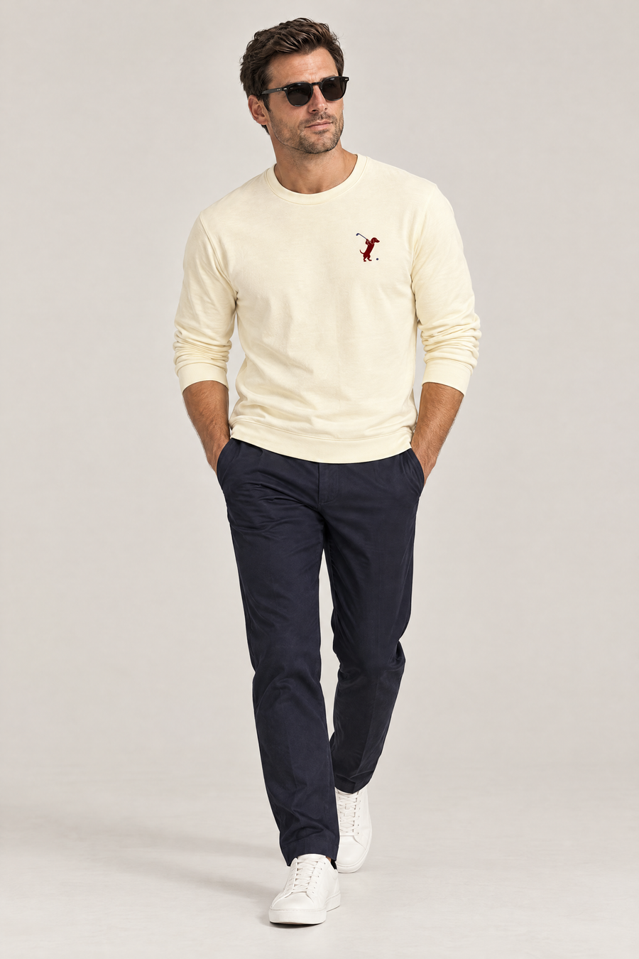 Man in sunglasses walking with hands in pockets wearing apricot Swing dachshund golf sweatshirt and dark navy trousers, full-body studio look. Color: Apricot