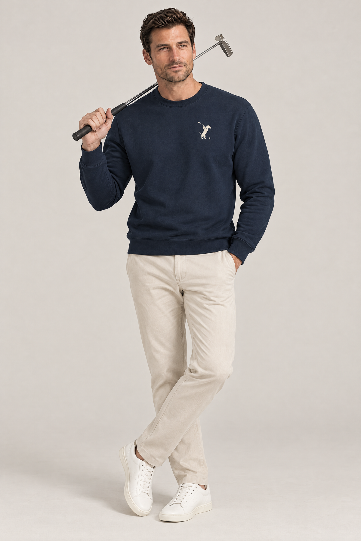 Smiling man with a putter resting on his shoulder wearing navy Swing dachshund golf sweatshirt and light beige trousers, full-body studio look. Color: Navy