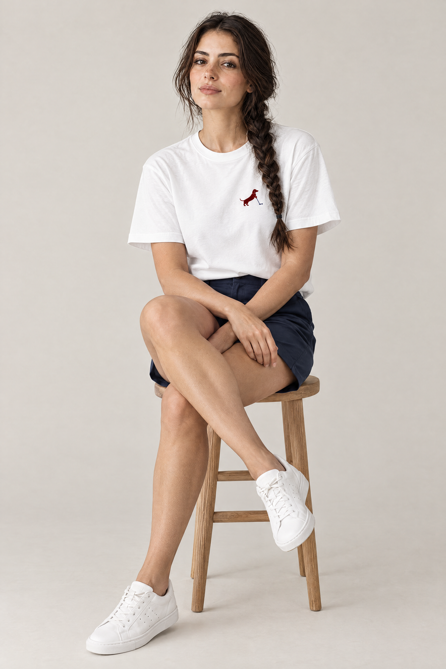 Woman sitting on a wooden stool wearing white Putting dachshund golf t-shirt with navy shorts. Color: White