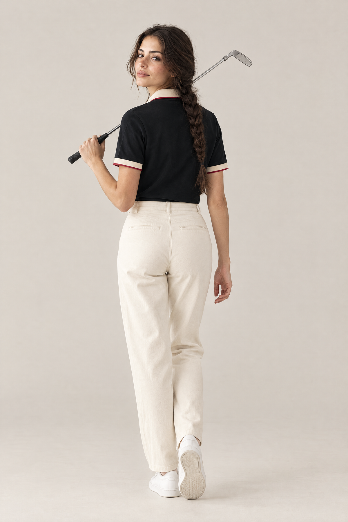 Woman walking away with a putter over her shoulder in the black Club Crest polo and cream trousers. Color: Black with Apricot