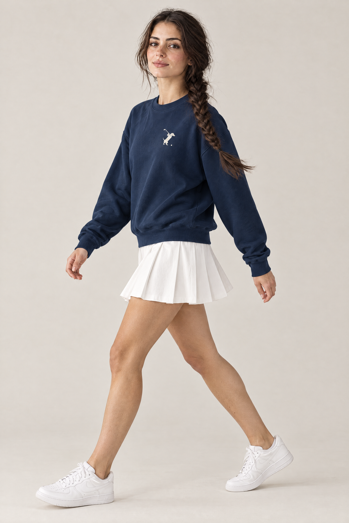 Woman walking in side view in navy Swing dachshund golf sweatshirt and white pleated tennis skirt. Color: Navy