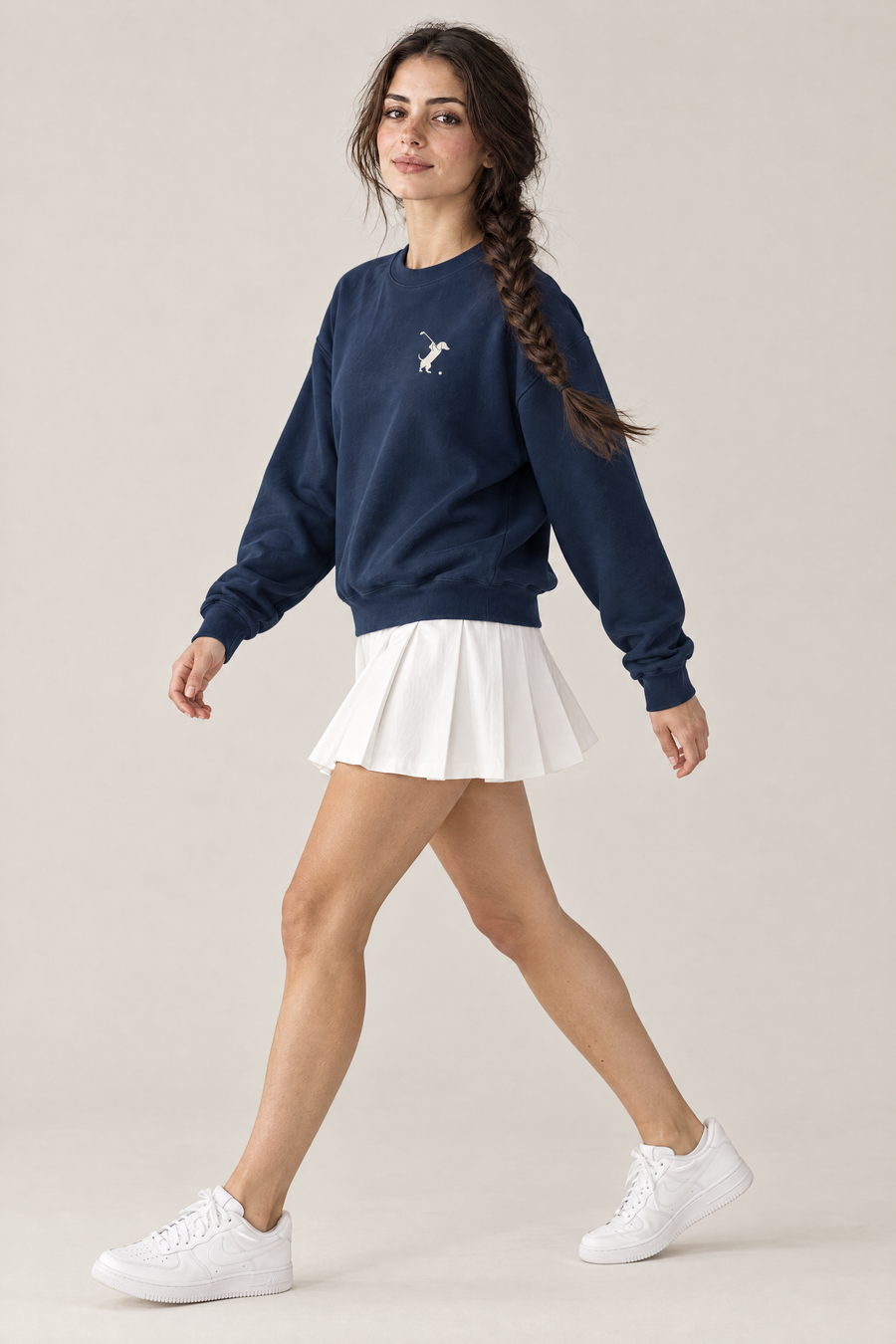 Woman walking in side view in navy Swing dachshund golf sweatshirt and white pleated tennis skirt. Color: Navy