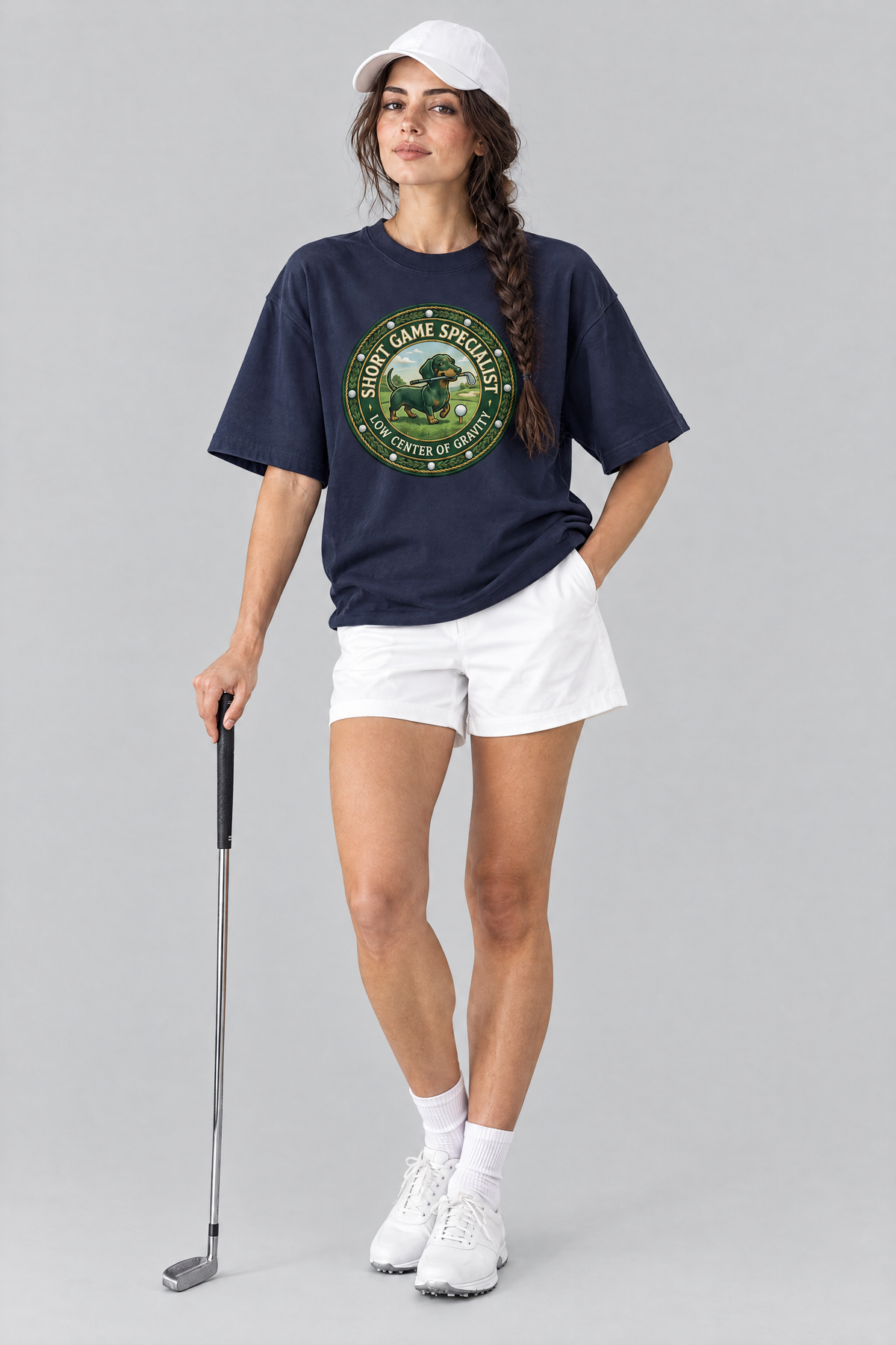 Woman wearing the Short Game Crest Dachshund Golf Tee in navy with white golf shorts and a white cap. Color: Navy