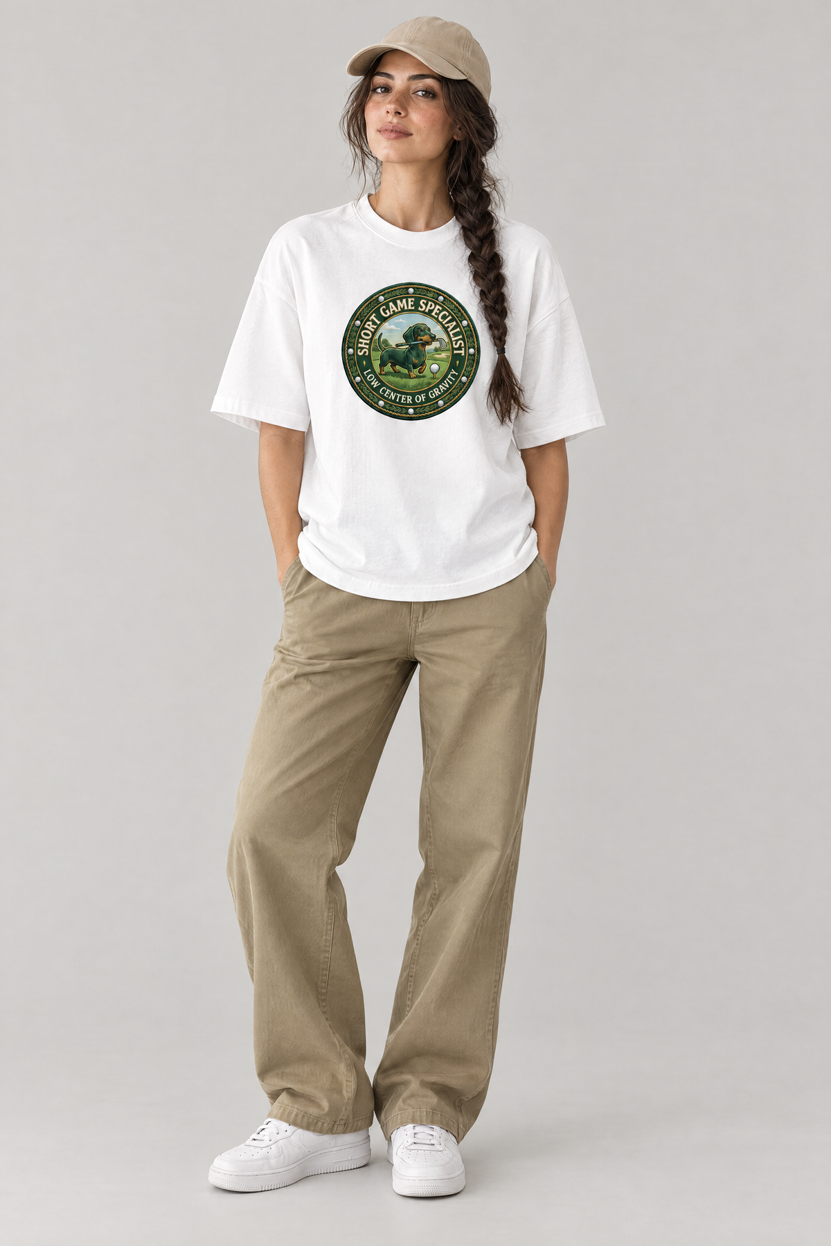 Woman wearing the Short Game Crest Dachshund Golf Tee in white with khaki trousers and a beige cap. Color: White