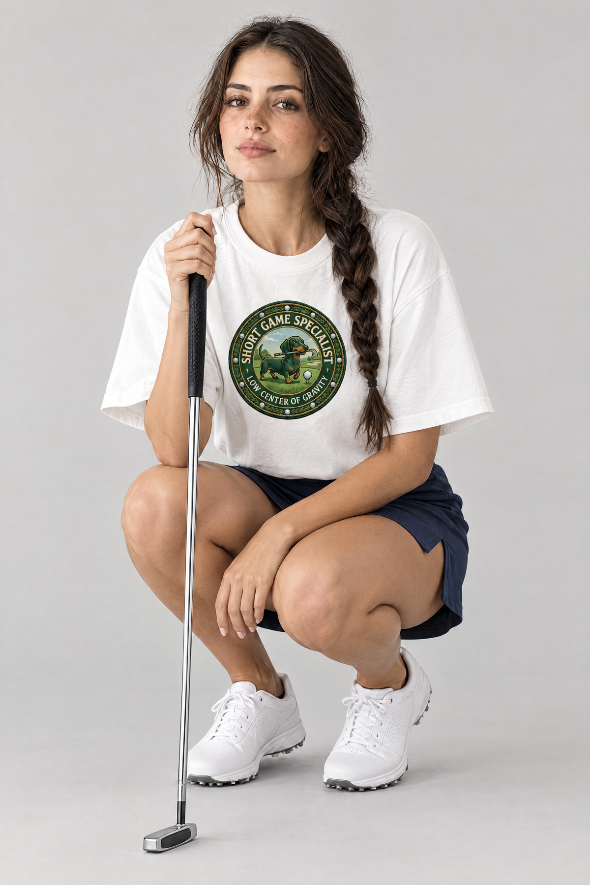 Woman lining up a putt in the white Short Game Crest Dachshund Golf Tee and a navy golf skort. Color: White