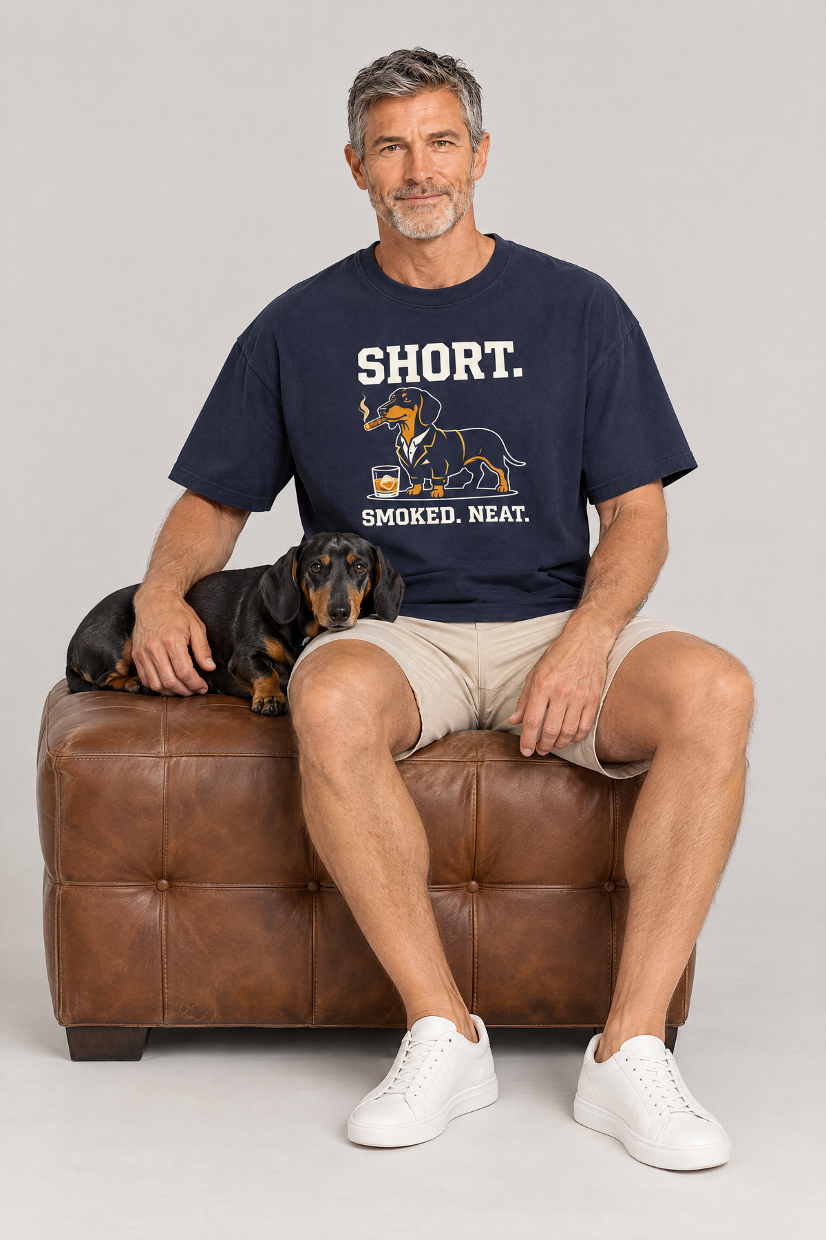 Man sitting on a leather ottoman with a black and tan dachshund in the navy Short Smoked Neat Dachshund Tee. Color: Navy